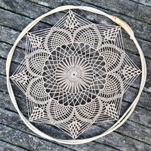 Puede incluir: Un gran atrapasueños de crochet beige con un patrón de estrella cuelga de un aro de madera. El atrapasueños está hecho con puntadas delicadas e intrincadas y tiene un círculo central con un círculo más pequeño en el centro.
