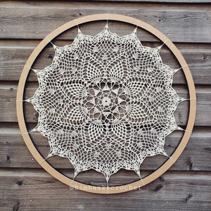 Puede incluir: Un encaje de ganchillo blanco con un dise&ntilde;o circular se muestra en un aro de madera. El encaje tiene un patr&oacute;n delicado e intrincado con una estrella central. El aro es marr&oacute;n y tiene un acabado de madera natural. El texto "@kimhaakcrochet" es visible en la parte inferior de la imagen.
