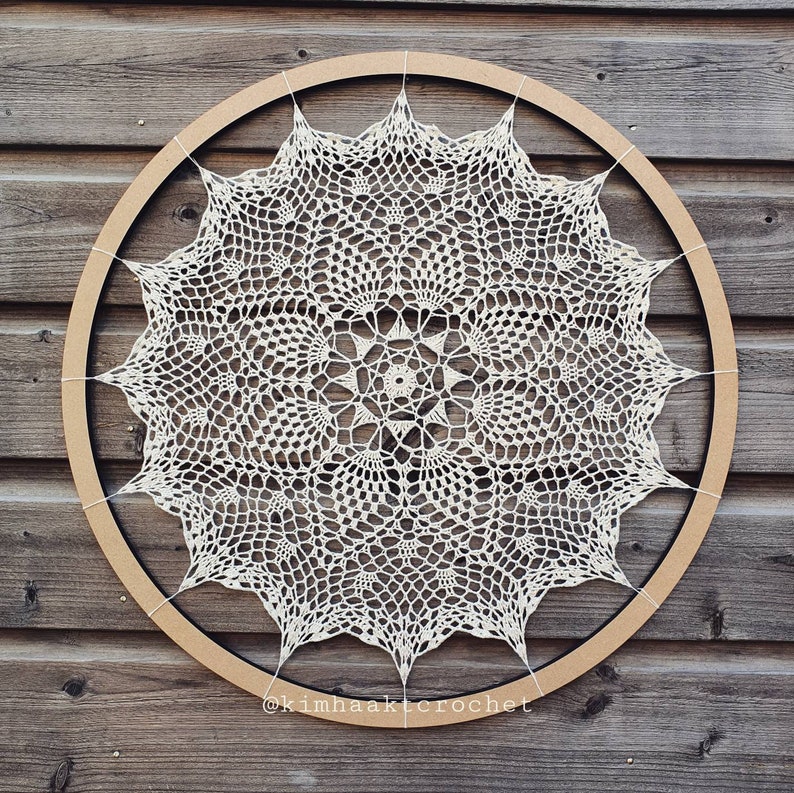Puede incluir: Un encaje de ganchillo blanco con un dise&ntilde;o circular se muestra en un aro de madera. El encaje tiene un patr&oacute;n delicado e intrincado con una estrella central. El aro es marr&oacute;n y tiene un acabado de madera natural. El texto "@kimhaakcrochet" es visible en la parte inferior de la imagen.