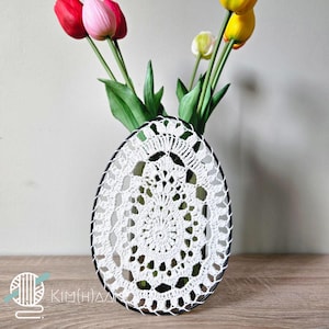 Op de afbeelding: Witte, ovaalvormige gehaakte vaas met een zwarte rand. De vaas bevat kleurrijke tulpen. De vaas heeft een ingewikkeld, opengewerkt ontwerp. De tekst "KIM(H)AA" is zichtbaar in de linker benedenhoek.