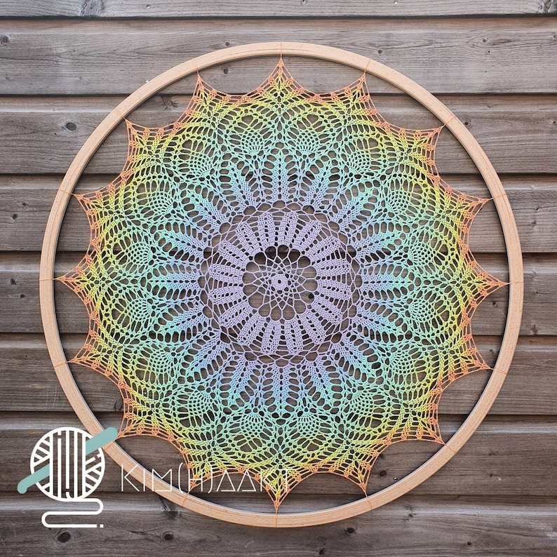 Puede incluir: Un gran mandala de crochet de colores arco&iacute;ris con un dise&ntilde;o delicado e intrincado. El mandala est&aacute; enmarcado en un aro de madera y cuelga sobre un fondo de veta de madera.