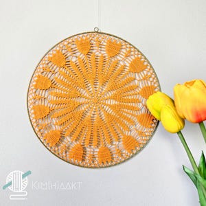 Op de afbeelding: Een ronde, oranje gehaakte wanddecoratie met een delicaat, kantpatroon. De decoratie is bevestigd aan een metalen ring en wordt getoond tegen een witte muur.