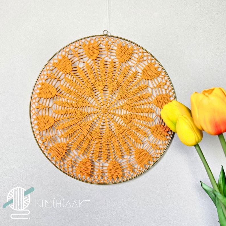 Op de afbeelding: Een ronde, oranje gehaakte wanddecoratie met een delicaat, kantpatroon. De decoratie is bevestigd aan een metalen ring en wordt getoond tegen een witte muur.
