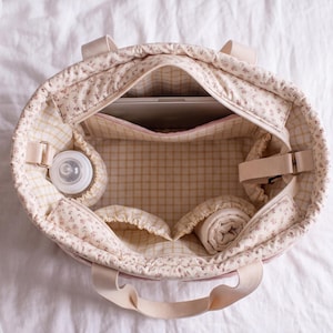 May include: A quilted tote bag with a cream and pink floral exterior and a yellow and white grid interior. The bag features multiple internal pockets, including a bottle holder, a laptop sleeve, and two fabric-lined compartments for storage.