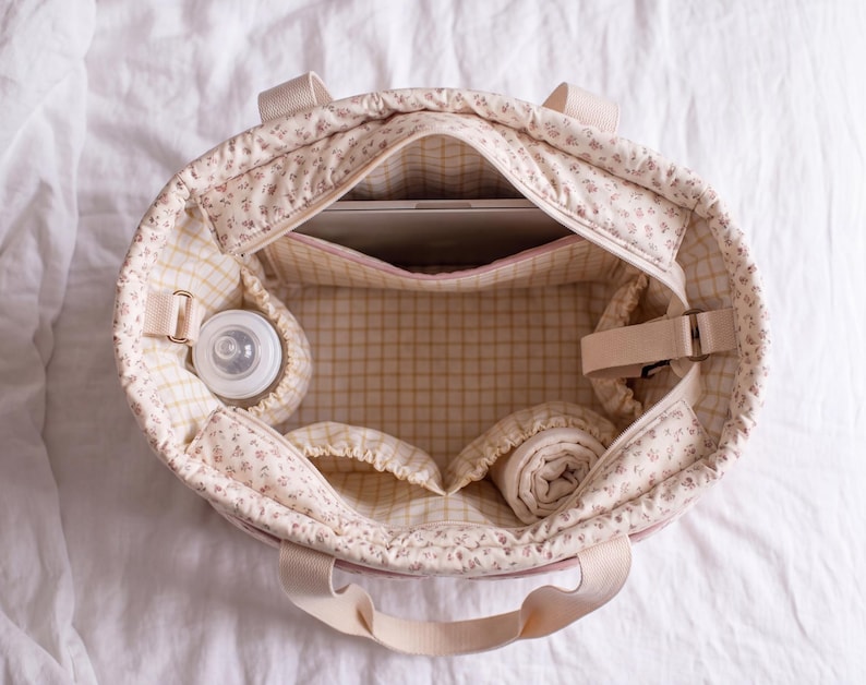 May include: A quilted tote bag with a cream and pink floral exterior and a yellow and white grid interior. The bag features multiple internal pockets, including a bottle holder, a laptop sleeve, and two fabric-lined compartments for storage.