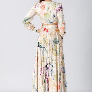 May include: A two-piece set consisting of a long-sleeved cropped top and a floor-length maxi skirt. Both pieces feature a cream-colored base with a watercolor-style floral print in shades of pink, purple, green, and blue, with long sleeves.