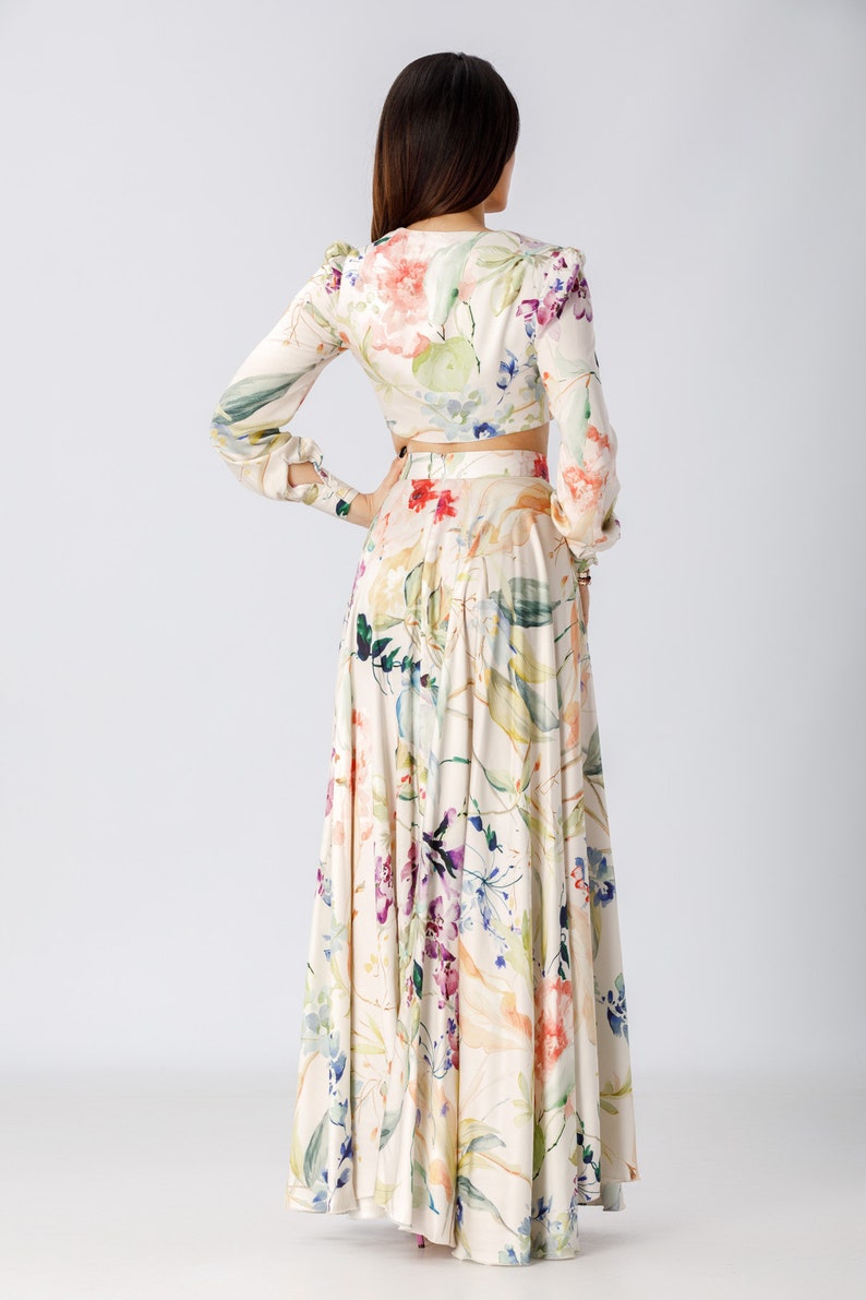 May include: A two-piece set consisting of a long-sleeved cropped top and a floor-length maxi skirt. Both pieces feature a cream-colored base with a watercolor-style floral print in shades of pink, purple, green, and blue, with long sleeves.
