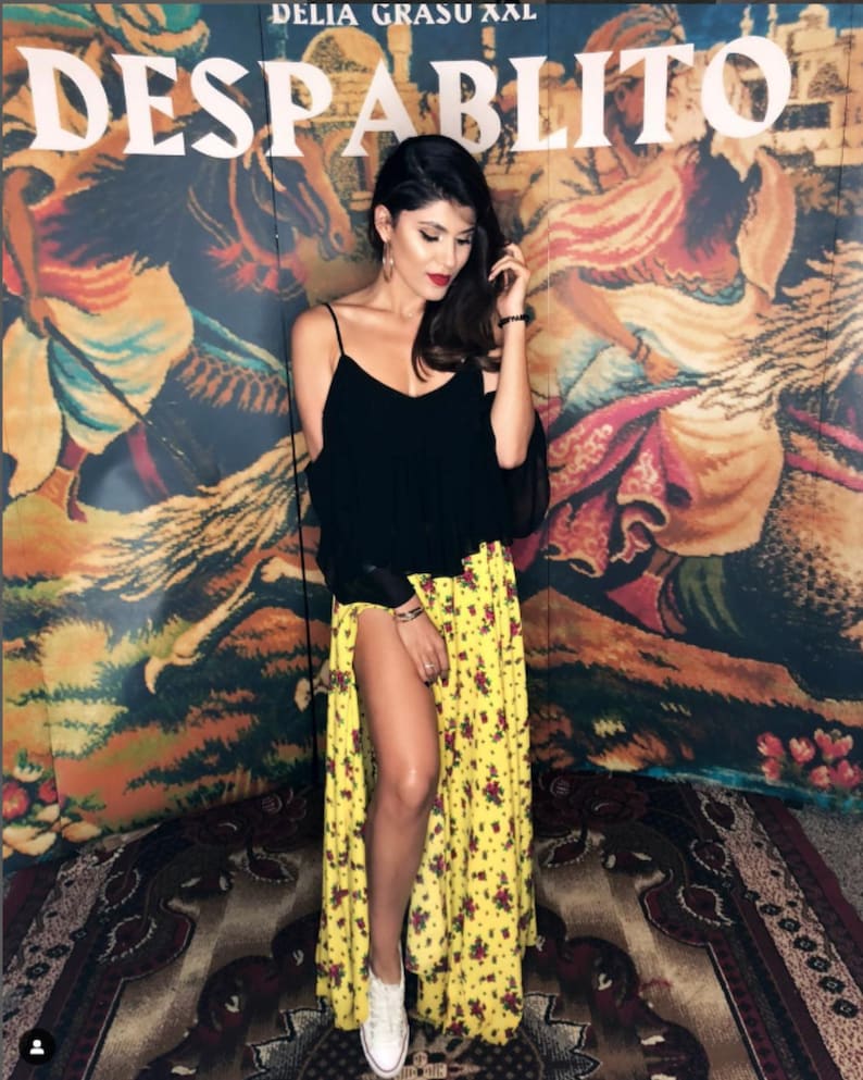 May include: A black sleeveless top with thin straps and a yellow floor-length skirt featuring a small red and green floral pattern and a high side slit, paired with white sneakers. The background displays the text DELIA GRASU XXL DESPABLITO.
