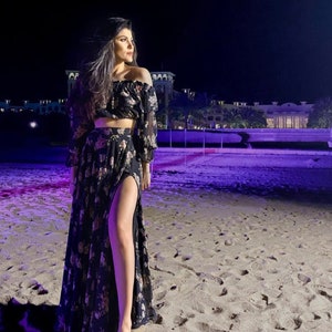 May include: A two-piece set consisting of an off-the-shoulder crop top with long sleeves and a matching floor-length skirt featuring a high side slit. The garment is made of black fabric with a repeating floral pattern in shades of pink and cream.