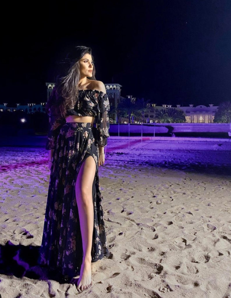 May include: A two-piece set consisting of an off-the-shoulder crop top with long sleeves and a matching floor-length skirt featuring a high side slit. The garment is made of black fabric with a repeating floral pattern in shades of pink and cream.