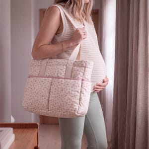 May include: A quilted tote bag featuring a cream-coloured base with a delicate pink floral pattern. The bag includes two sturdy shoulder straps, a rectangular shape, and two exterior front pockets with a solid pink trim along the top edge.