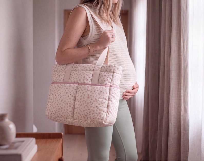 May include: A quilted tote bag featuring a cream-coloured base with a delicate pink floral pattern. The bag includes two sturdy shoulder straps, a rectangular shape, and two exterior front pockets with a solid pink trim along the top edge.