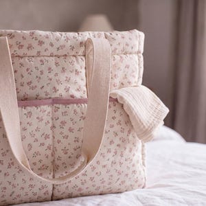 May include: A quilted tote bag featuring a cream-coloured fabric with a small pink floral pattern. The bag includes two wide, light beige fabric handles and a horizontal pocket across the front with a solid pink trim along the top edge.