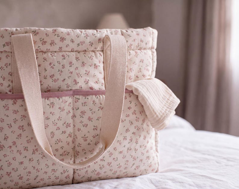 May include: A quilted tote bag featuring a cream-coloured fabric with a small pink floral pattern. The bag includes two wide, light beige fabric handles and a horizontal pocket across the front with a solid pink trim along the top edge.