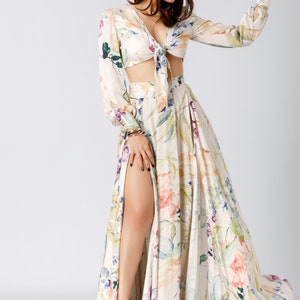 May include: A two-piece set featuring a long-sleeved crop top with a front tie closure and a matching floor-length maxi skirt with a high side slit. Both pieces are made of a cream-colored fabric with a multi-colored floral print in shades of pink, blue, and green.