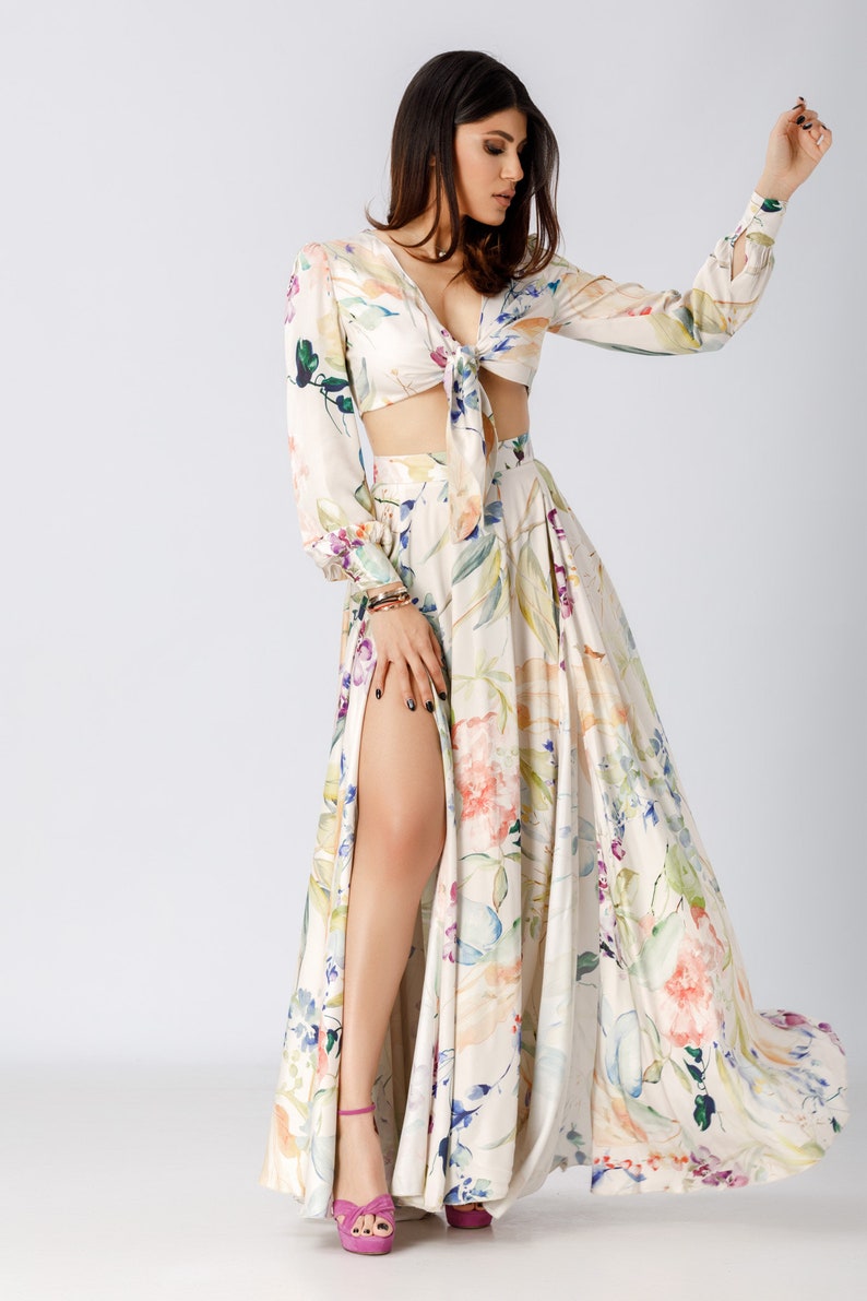 May include: A two-piece set featuring a long-sleeved crop top with a front tie closure and a matching floor-length maxi skirt with a high side slit. Both pieces are made of a cream-colored fabric with a multi-colored floral print in shades of pink, blue, and green.