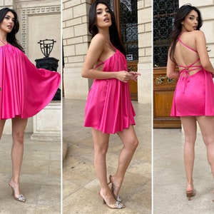 May include: A vibrant hot pink mini dress featuring a gathered halter neckline, a loose flowing silhouette, and thin spaghetti straps that crisscross across the open back. The garment is made of a smooth, satin-like fabric with a subtle sheen.