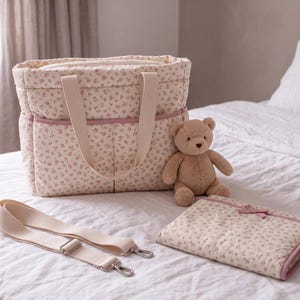 May include: A quilted nappy bag with a cream and pink floral pattern, featuring two top handles and a zip closure. Included are a detachable cream shoulder strap with metal hardware, a matching folded changing mat, and a small tan teddy bear.