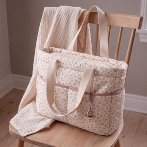 May include: A quilted tote bag featuring a cream-coloured fabric with a small-scale pink floral pattern. The bag includes two cream-coloured fabric handles, two exterior front pockets with pink trim, and a dummy attached to a small strap.