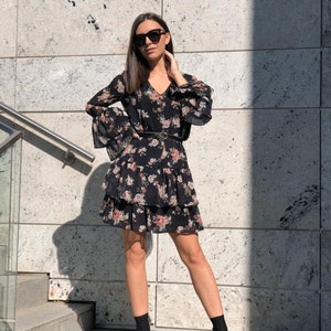 May include: A black floral dress with a tiered skirt and long sleeves. The dress has a V-neckline and a black belt. The model is wearing black ankle boots.