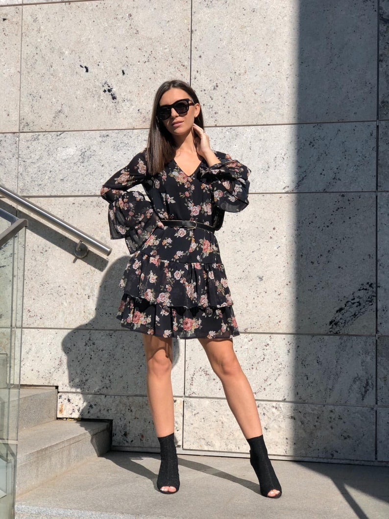 May include: A black floral dress with a tiered skirt and long sleeves. The dress has a V-neckline and a black belt. The model is wearing black ankle boots.