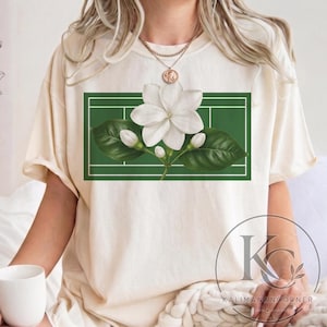 May include: A cream-colored short-sleeve t-shirt featuring a rectangular graphic on the chest. The graphic displays a white jasmine flower with green leaves centered over a dark green tennis court design with white boundary lines.