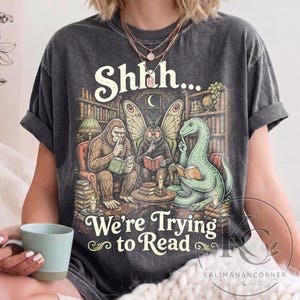 May include: A charcoal gray short-sleeve t-shirt featuring a graphic of a sasquatch, a moth creature, and a dinosaur sitting in a library reading books with the text Shhh... We're Trying to Read printed in a decorative cream-colored font.