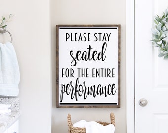 May include: A white bathroom sign with black text that reads "Please stay seated for the entire performance". The sign is hanging on a white wall above a wicker laundry basket.