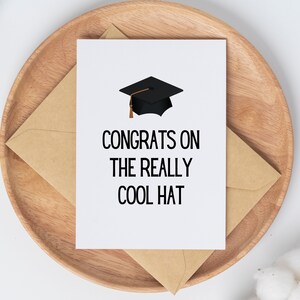 以下が含まれることがあります： 「Congrats on the really cool hat」という黒い文字が書かれた白いカード。文字の上に、金色の房飾りがついた黒い卒業帽が描かれている。