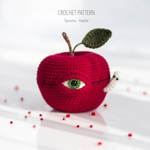 Könnte beinhalten: Ein gehäkelter roter Apfel mit einem aufgestickten Auge und einem weißen Wurm, der aus der Seite kommt. Der Apfel hat ein grünes Blatt und einen Stiel. Der Text "CROCHET PATTERN Spooky Apple" steht oben.