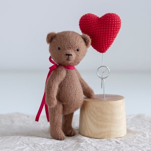 Może przedstawiać: Brązowy miś z czerwoną wstążką wokół szyi stoi obok czerwonego serduszka wykonanego na szydełku na drewnianym stojaku.