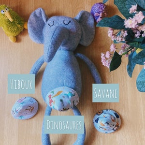 Peut inclure: Un éléphant en peluche gris avec une couche à motifs bleus et blancs. La couche a un imprimé de dinosaures. L'éléphant est couché sur une surface en bois avec trois petits chapeaux à proximité. Les chapeaux sont étiquetés "Hiboux", "Savane" et "Dinosaures".