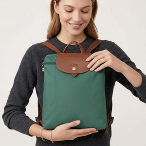 May include: Green backpack with a brown leather flap and handle. The backpack has brown shoulder straps and a gold zip. The bag is held by a person wearing a dark grey jumper.