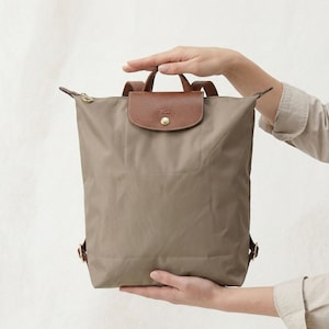 May include: A taupe backpack with a brown leather flap and handle. The backpack has gold-colored zippers and hardware. The bag is held up by a person's hands.
