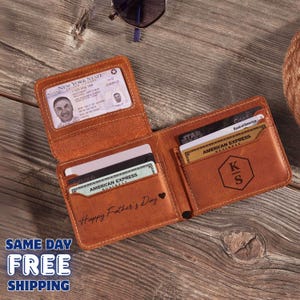 May include: Open brown leather wallet with multiple card slots, holding credit cards, a driver's license, and a personalized monogram. The wallet is embossed with "Happy Father's Day". Sunglasses are visible in the background.