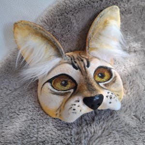 Puede incluir: Una máscara de gato hecha a mano, en tonos beige y marrón, con grandes orejas erguidas y una nariz negra. Los ojos están detallados con una malla amarilla, y la máscara tiene un aspecto realista y peludo. Diseñada para cosplay o disfraces.