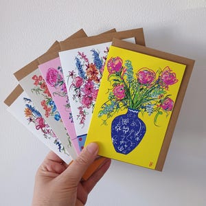 Könnte beinhalten: Eine Hand hält einen Stapel Grußkarten mit floralen Designs. Die Karten zeigen farbenfrohe Illustrationen von Blumen in Vasen und Sträußen. Die Karten haben eine Rückseite aus Kraftpapier und befinden sich auf weißem Hintergrund.