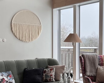 Peut inclure: Un salon avec de grandes fenêtres donnant sur une forêt enneigée. La pièce comprend un canapé vert avec des coussins, une table basse en bois, deux fauteuils gris, une lampe sur pied et une tapisserie murale en macramé.