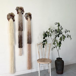 May include: Three ceramic wall hangings with long, flowing strands of yarn in white, brown, and beige. The hangings are displayed on a white wall with a wooden chair and a potted plant in the foreground.