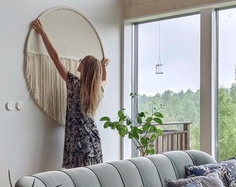 May include: A woman hangs a large, round, white macrame wall hanging with fringe on a white wall. The wall hanging is in the center of the image. The woman is wearing a black and white patterned dress. The wall hanging is in a living room with a green sofa, pillows, and plants.