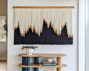 May include: A wooden console table with two shelves and a white wall behind it. The table has four blue painted legs and a natural wood top. A black and white yarn wall hanging is above the table.