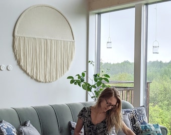 May include: A woman sits on a green sofa with a cat in her lap. The sofa has several pillows with floral patterns. A large, white, circular wall hanging with fringe is on the wall behind the sofa. A small potted plant is on the table next to the sofa.