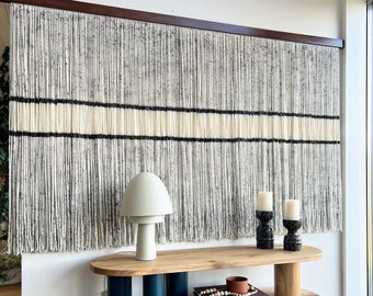 May include: A wooden console table with a light wood top and teal blue legs. The table has two shelves and is decorated with a white ceramic lamp, two black candle holders, and a stack of books. The wall behind the table is decorated with a macrame wall hanging with black and white yarn.