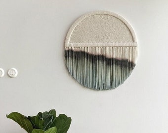 May include: A round wall hanging with a white and beige woven center and a fringe of blue and green yarn. The hanging is on a white wall with a potted plant and a basket with pillows in the foreground.