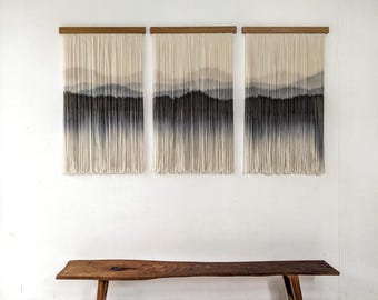 May include: Three wall hangings with a landscape design. Each piece features a wooden rod at the top, with cream-colored strands that fade into shades of gray and black, creating a mountain silhouette. A wooden bench sits below.