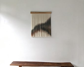 May include: A wall hanging featuring a wooden top bar with vertical cream-colored yarn strands. A dark brown and black gradient pattern is dyed into the lower section of the yarn, creating a sweeping, curved shape across the fiber strands.