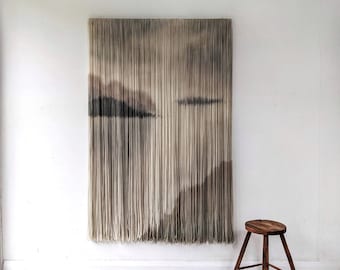 May include: A large, rectangular wall hanging with a landscape design. The artwork features vertical strands in shades of beige and brown, creating a textured effect. A small, wooden stool with a round seat and three legs is positioned to the right.