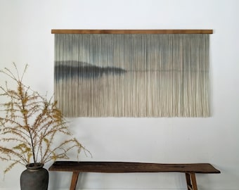 May include: A large wall hanging with a landscape design, featuring a wooden frame and vertical strands in shades of gray and beige. A dark wooden bench and a vase with dried branches are in front of a white wall.
