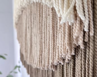 May include: Close-up of a textured wall hanging with layers of off-white, beige, and brown yarn. The design features a combination of fringed and twisted yarn, creating a tactile and layered effect. This handcrafted piece adds a touch of bohemian style.