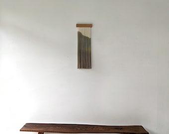 May include: A wall hanging featuring a rectangular wooden top bar with vertical, multi-colored fiber strands in shades of cream, olive green, and dark brown, suspended above a long, dark brown wooden bench with a natural live edge and four legs.